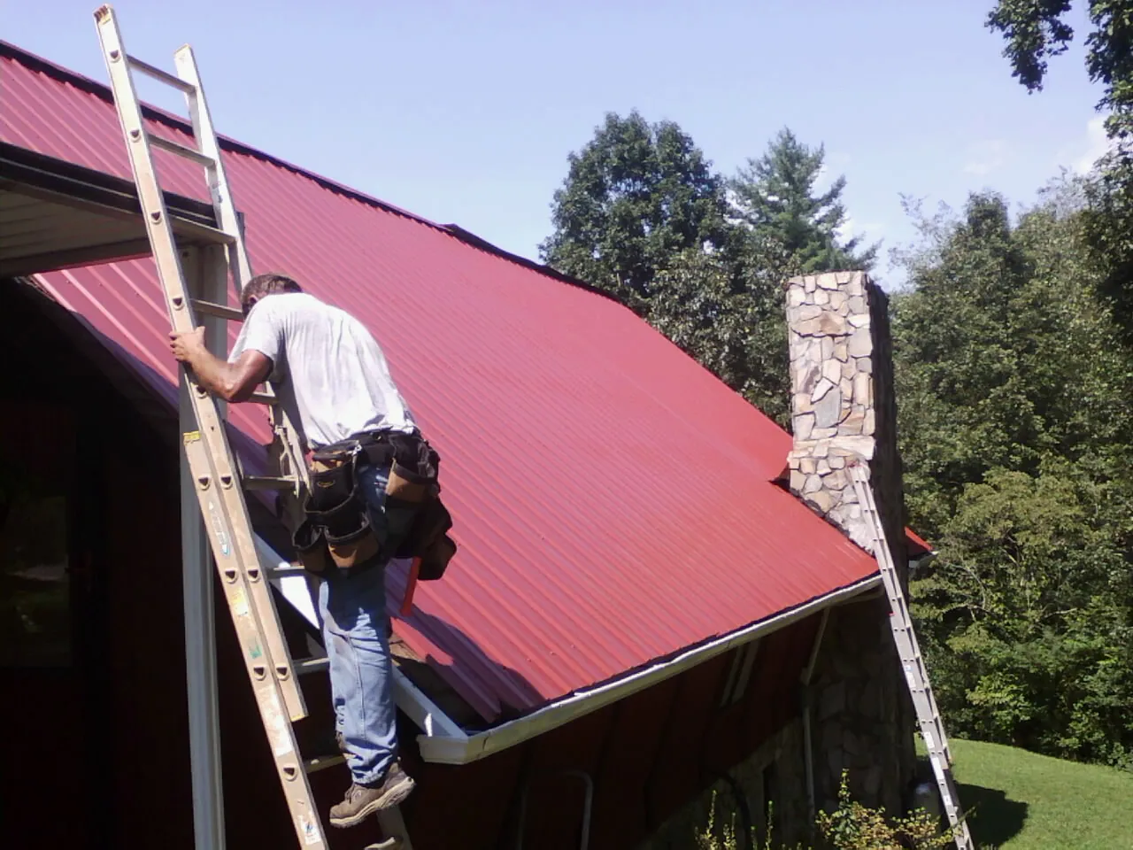 Roofer on a ladder beside a red metal roof and stone chimney — Soffit & Fascia Repair by Roofing Brothers in Francitas